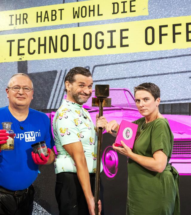 Das Bild zeigt drei Personen, die nebeneinander stehen. Im Hintergrund ist ein grafisches Muster mit der Aufschrift "IHR HABT WOHL DIE TECHNOLOGIE OFFEN" in gelben Buchstaben auf einem dunkelgrauen Untergrund. Die erste Person links trägt ein blaues T-Shirt mit der Aufschrift "upTuni Wien" und hält eine Glasflasche sowie ein schwarzes Behältnis in den Händen. Sie hat eine Brille auf und trägt rote Handschuhe.   In der Mitte steht eine zweite Person in einem hellen Hemd mit einem bunten Muster aus kleinen Figuren. Diese Person hat eine verspielte Miene und hält einen langen Stock.   Die dritte Person auf der rechten Seite trägt ein grünes Oberteil und sieht ernst aus. Sie hält ein pinkes Notizbuch oder ein ähnliches Objekt in der Hand und hat eine kurze Frisur. Im Hintergrund sind farbenfrohe grafische Elemente, darunter ein pinkes Auto und eine cartoonartige Illustration eines Tieres. Die gesamte Szenerie hat einen modernen und dynamischen Charakter.