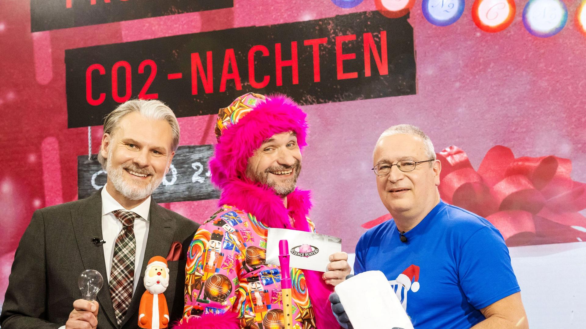 Das Bild zeigt drei Männer, die vor einem festlich dekorierten Hintergrund stehen. An der Wand sind mehrere Weihnachtslichter und ein großes Banner mit der Aufschrift "FROHE CO2-NACHTEN" zu sehen. Der erste Mann, links im Anzug, hält einen Weihnachtsmann in der Hand und eine Glühbirne. Er hat kurzes graues Haar und einen Bart. Der zweite Mann, in der Mitte, trägt ein buntes, auffälliges Outfit mit einem pinken Pelzkragen. Er hält eine Pfeife und ein Schild mit der Aufschrift "Science Busters". Der dritte Mann, rechts, trägt ein blaues T-Shirt und eine Schutzbrille. In seiner Hand hält er einen Block Eis. Alle drei Männer scheinen fröhlich zu sein und lächeln in die Kamera. Der Hintergrund zeigt eine festliche Atmosphäre, die zum Thema Weihnachten passt.