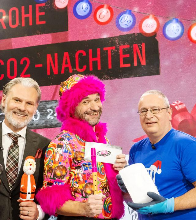 Das Bild zeigt drei Männer, die vor einem festlich dekorierten Hintergrund stehen. An der Wand sind mehrere Weihnachtslichter und ein großes Banner mit der Aufschrift "FROHE CO2-NACHTEN" zu sehen. Der erste Mann, links im Anzug, hält einen Weihnachtsmann in der Hand und eine Glühbirne. Er hat kurzes graues Haar und einen Bart. Der zweite Mann, in der Mitte, trägt ein buntes, auffälliges Outfit mit einem pinken Pelzkragen. Er hält eine Pfeife und ein Schild mit der Aufschrift "Science Busters". Der dritte Mann, rechts, trägt ein blaues T-Shirt und eine Schutzbrille. In seiner Hand hält er einen Block Eis. Alle drei Männer scheinen fröhlich zu sein und lächeln in die Kamera. Der Hintergrund zeigt eine festliche Atmosphäre, die zum Thema Weihnachten passt.