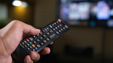 Großaufnahme einer Hand die eine Fernbedienung hält um Tasten zu drücken. Im Hintergrund unscharf ein Fernseher.