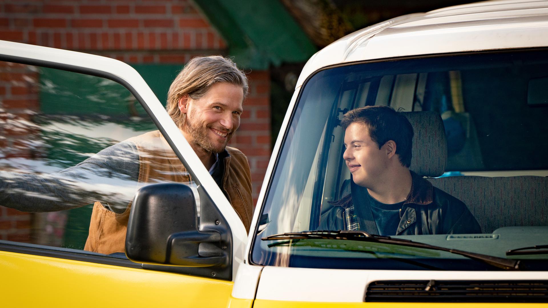 Alex (Sebastian Ströbel) steht vor der offenen Beifahrertür seines Autos und lacht seinen Sohn Felix an, der bereits auf dem Beifahrersitz sitzt.