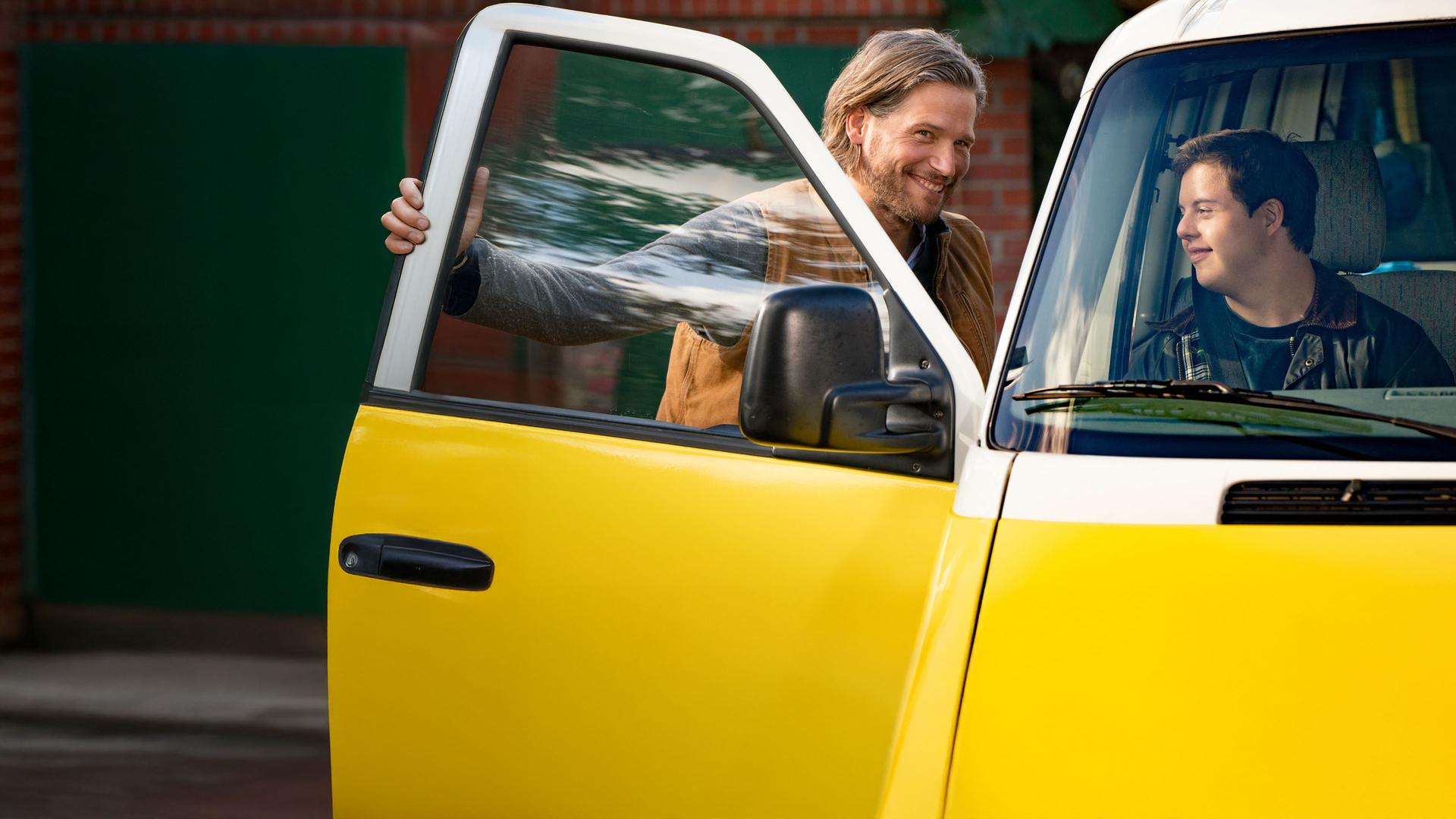Alex (Sebastian Ströbel) steht vor der offenen Beifahrertür seines Autos und lacht seinen Sohn Felix an, der bereits auf dem Beifahrersitz sitzt.