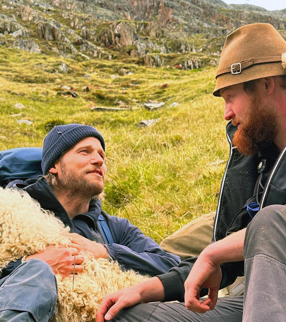 Sebastian Ströbel liegt auf einer Wiese und hat ein Schaf im Arm. Der Schäfer Manuel Götsch sitzt daneben.