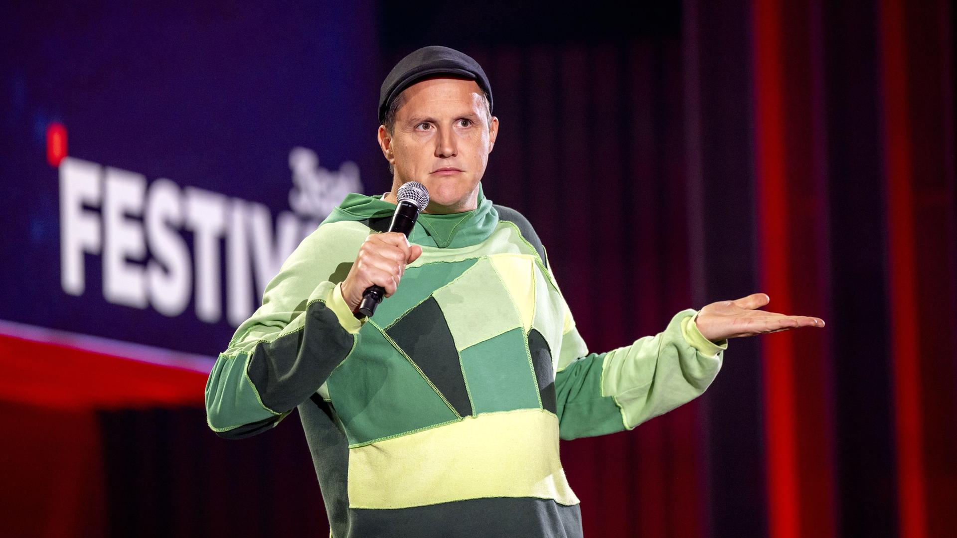 Sebastian23 steht in der Mitte des Bildes. Seine linke Hand streckt er mit der Handinnenfläche nach oben vor sich aus. In der rechten Hand hält er ein Mikrofon. Er trägt einen grünen Pullover und eine Mütze.