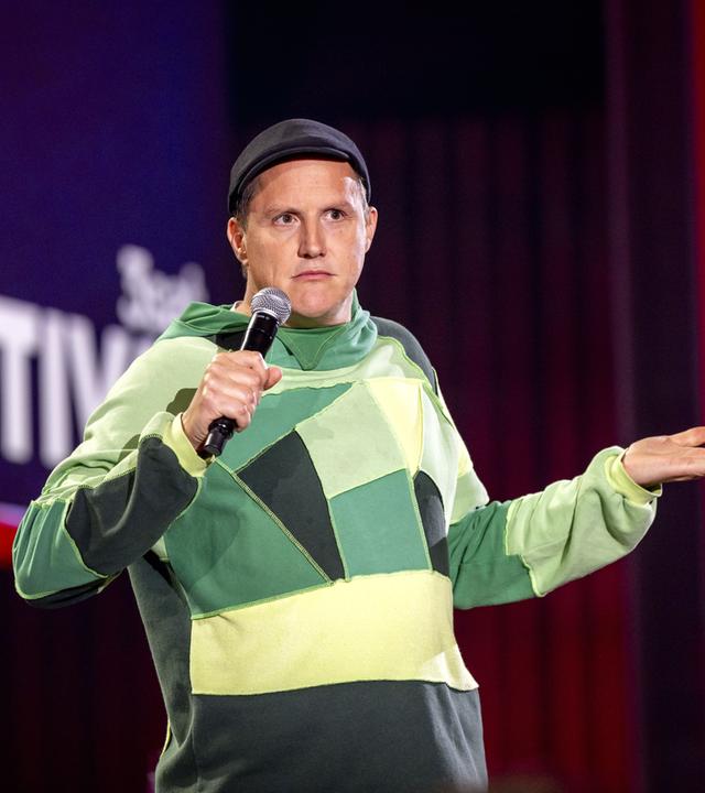 Sebastian23 steht in der Mitte des Bildes. Seine linke Hand streckt er mit der Handinnenfläche nach oben vor sich aus. In der rechten Hand hält er ein Mikrofon. Er trägt einen grünen Pullover und eine Mütze.