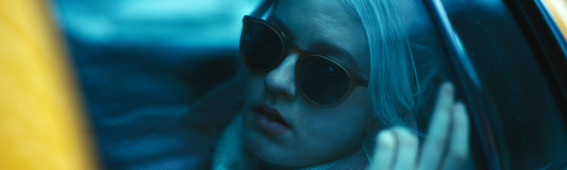 "See for Me - Der unsichtbare Feind": Nahaufnahme: Blick durch blau getönte Fensterscheibe durch die Taxiseitentür auf Sophie (Sklyer Davenport), die mit Sonnenbrille auf Rückbank sitzt.