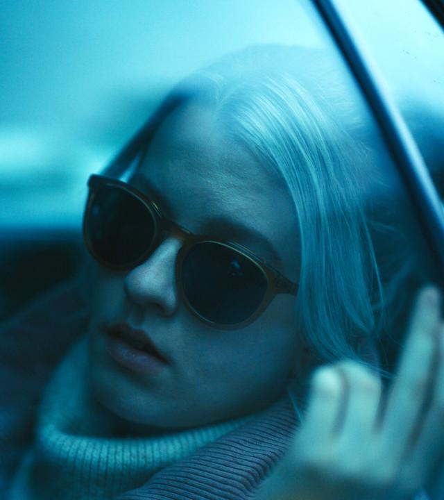 "See for Me - Der unsichtbare Feind": Nahaufnahme: Blick durch blau getönte Fensterscheibe durch die Taxiseitentür auf Sophie (Sklyer Davenport), die mit Sonnenbrille auf Rückbank sitzt.