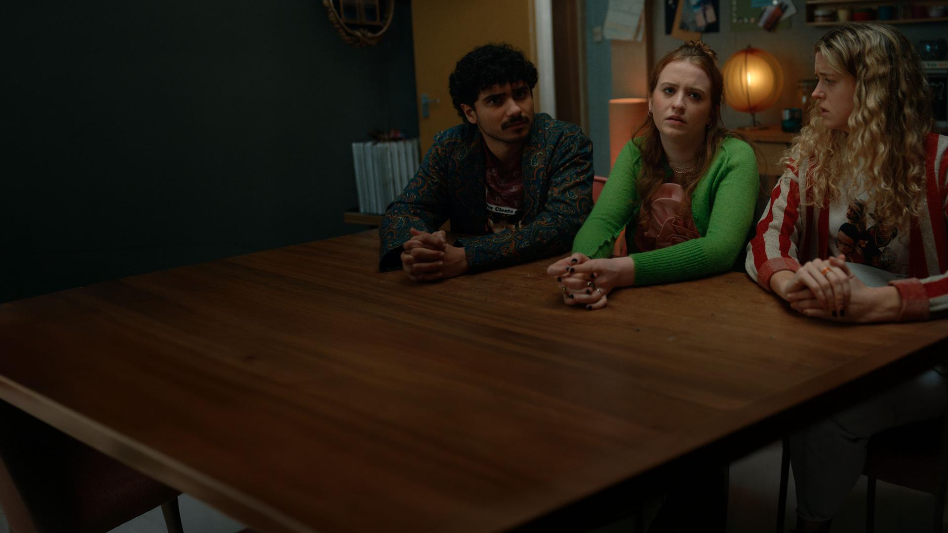 "Extraordinary - Sei der Daddy, den die Welt braucht": Kash (Bilal Hasna) sitzt neben Jen (Máiréad Tyers) und Carry (Sofia Oxenham) am Holztisch in der Küche. Jen wirkt verzweifelt. Sie hat die Hände auf dem Tisch verschränkt. Carry schaut sie verdutzt an. Im Hintergrund brennt eine kleine Tischlampe.