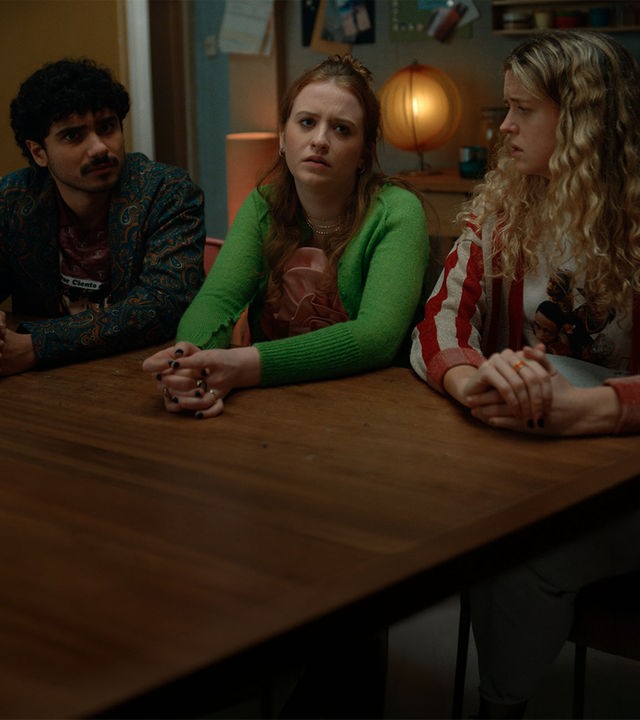 "Extraordinary - Sei der Daddy, den die Welt braucht": Kash (Bilal Hasna) sitzt neben Jen (Máiréad Tyers) und Carry (Sofia Oxenham) am Holztisch in der Küche. Jen wirkt verzweifelt. Sie hat die Hände auf dem Tisch verschränkt. Carry schaut sie verdutzt an. Im Hintergrund brennt eine kleine Tischlampe.