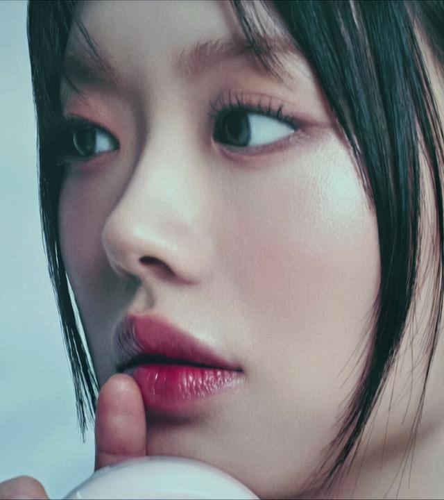 Das Bild zeigt ein großes, nahes Porträt eines Gesichts einer Frau mit glatter, makelloser Haut. Ihr Blick ist direkt nach vorne gerichtet, und ihre Lippen sind leicht geöffnet, als ob sie einen Finger an den Mund legt, was einen nachdenklichen oder geheimnisvollen Ausdruck vermittelt. Die Haare sind dunkel und in einem geraden Schnitt styled, und die Haut hat einen sichtbaren Glanz. Der Hintergrund ist unscharf und in einem hellen Farbton gehalten, was dem Bild eine sanfte und beruhigende Atmosphäre verleiht. Es scheint sich um eine Werbung oder ein Plakat für ein Schönheitsprodukt zu handeln, da das Motiv stark an die K-Beauty-Ästhetik erinnert.