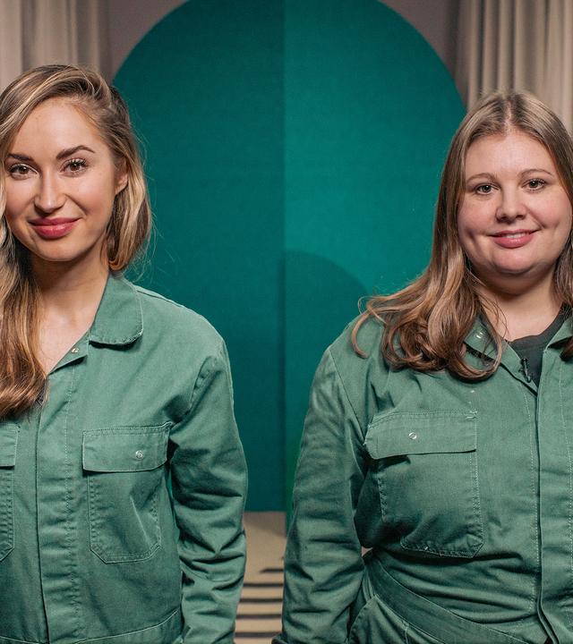 Zwei Frauen blicken in die Kamera. Sie tragen beide den selben grünen Overall.