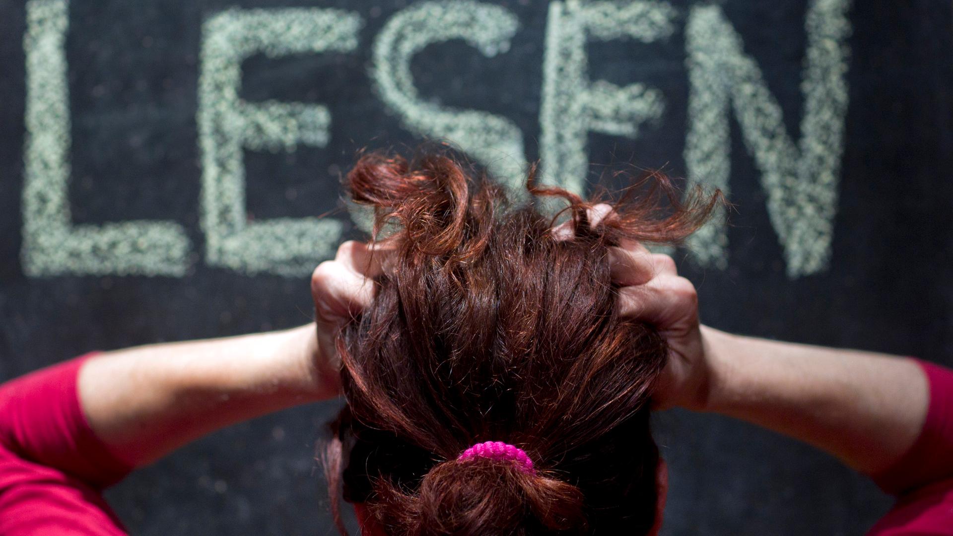 Eine Frau ist von hinten zu sehe, rauft sich die Haare beim Blick auf eine Tafel, auf der das Wort LESEN steht.