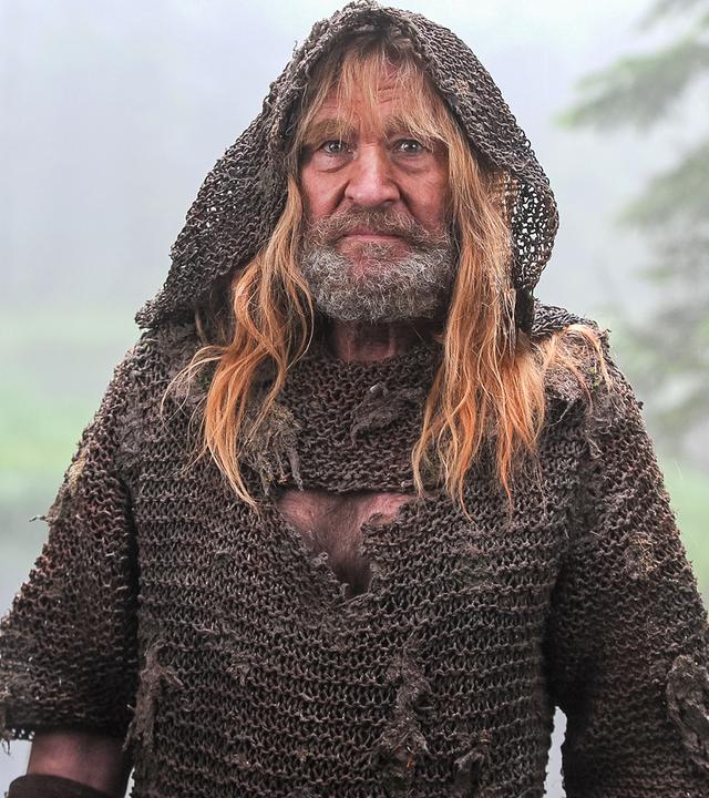 Vor dem Hintergrund eines Waldsees, der in eine Wiese ausläuft steht der Eisenhans (Michael Mendl). Er trägt Bart, eine weite Kapuze über dem Kopf, hat lange, blonde Haare und trägt ein schmutzig-braun wirkendes Gewand.