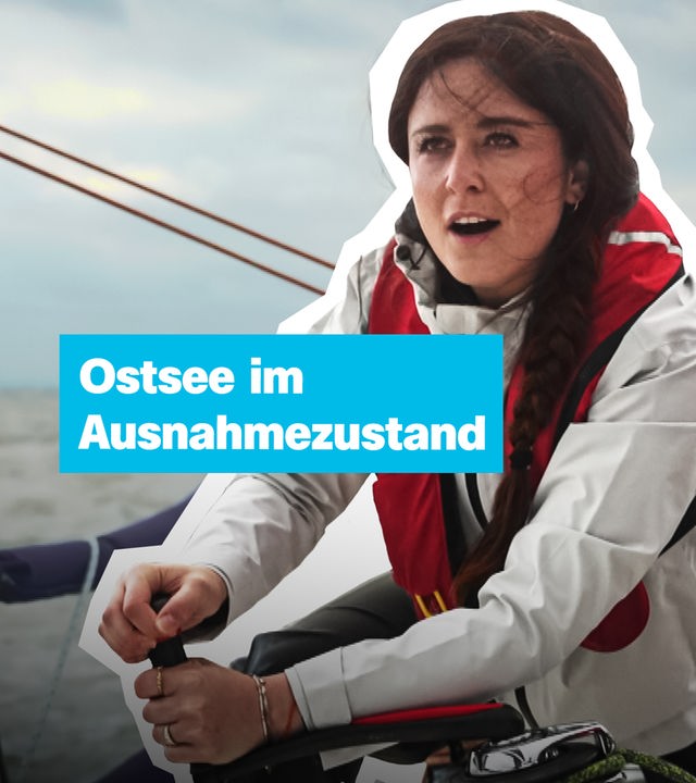 Auf einem Segelboot bei stürmischer See sitzt eine junge Frau in heller Jacke und roter Rettungsweste und kurbelt an einer Kurbel. 