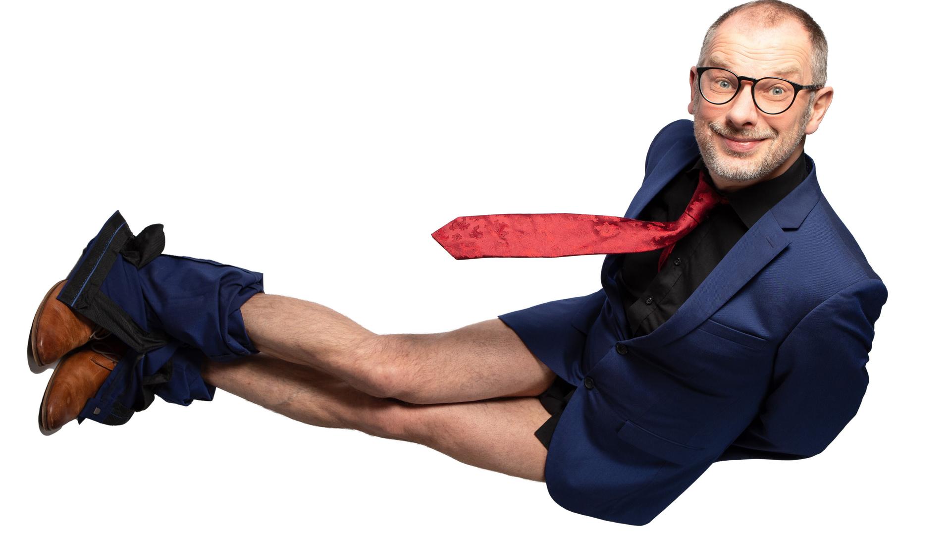 Das Bild zeigt einen Mann, der in einer unkonventionellen Pose auf einer weißen Hintergrundfläche steht. Er trägt einen blauen Anzug mit einem schwarzen Hemd und einer roten Krawatte. Seine Unterhose ist sichtbar, da seine Hose bis zu den Knien heruntergezogen ist. Der Mann hat kurze Haare, eine Brille und ein breites Lächeln auf dem Gesicht, was ihm einen freundlichen Ausdruck verleiht. Seine braunen Schuhe sind gut sichtbar. Die Komposition und der Kontrast zwischen dem Anzug und der weißen Hintergrundfläche betonen den humorvollen Charakter der Pose.