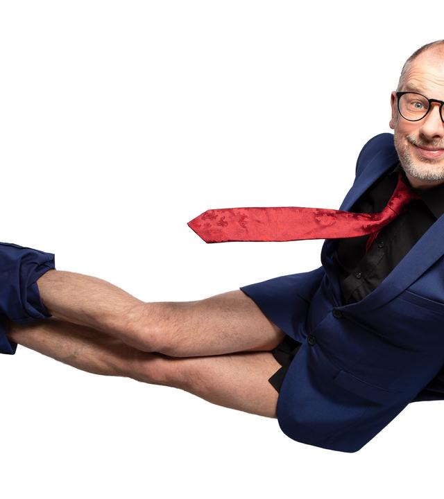 Das Bild zeigt einen Mann, der in einer unkonventionellen Pose auf einer weißen Hintergrundfläche steht. Er trägt einen blauen Anzug mit einem schwarzen Hemd und einer roten Krawatte. Seine Unterhose ist sichtbar, da seine Hose bis zu den Knien heruntergezogen ist. Der Mann hat kurze Haare, eine Brille und ein breites Lächeln auf dem Gesicht, was ihm einen freundlichen Ausdruck verleiht. Seine braunen Schuhe sind gut sichtbar. Die Komposition und der Kontrast zwischen dem Anzug und der weißen Hintergrundfläche betonen den humorvollen Charakter der Pose.