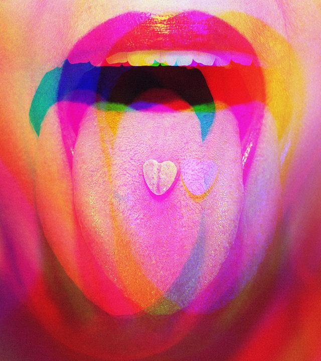 Nahaufnahme eines geöffneten Mundes mit ausgestreckter Zunge; darauf liegt eine kleine herzförmige Pille, überlagert von bunten, verschwommenen Farbeffekten.