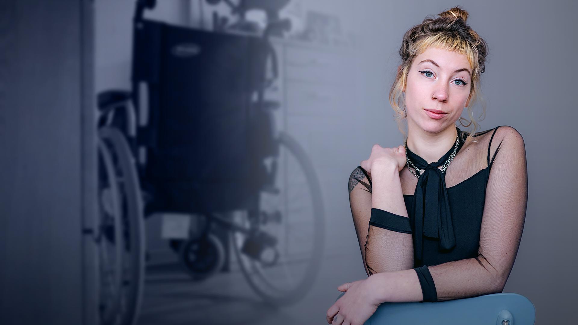 Pamina ist im Vordergrund zu sehen. Sie hat eine Hand auf einer Stuhllehne aufgelegt und die andere Hand hält sie auf ihre linke Schulter. Im Hintergrund ist ein Rollstuhl zu sehen. 