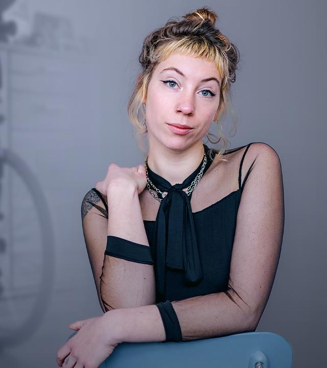 Pamina ist im Vordergrund zu sehen. Sie hat eine Hand auf einer Stuhllehne aufgelegt und die andere Hand hält sie auf ihre linke Schulter. Im Hintergrund ist ein Rollstuhl zu sehen. 