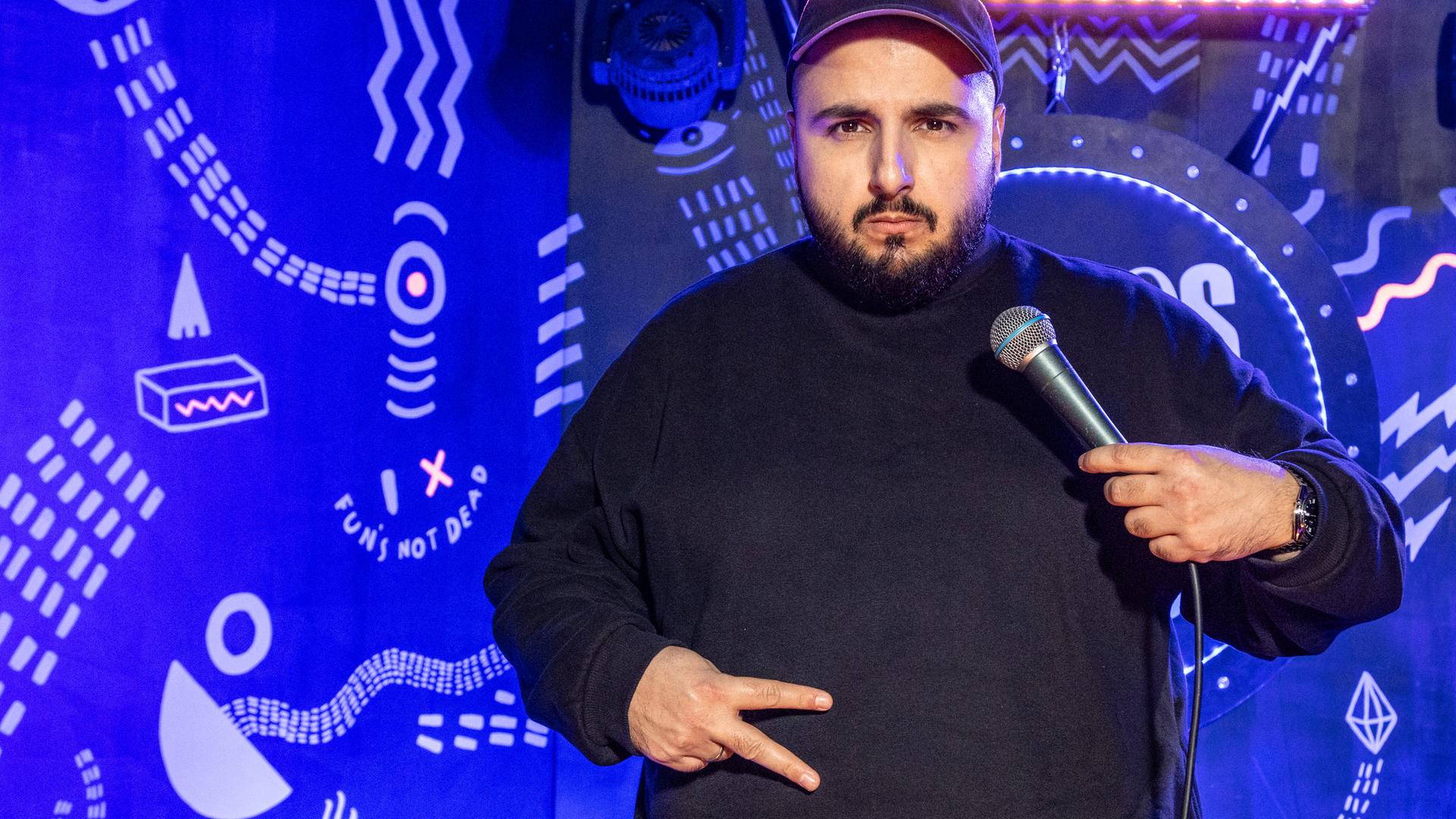 Sezer Oglu steht auf der Bühne und zeigt mit seiner rechten Hand das Peace Zeichen vor seinem Bauch. In der linken Hand hat er ein Mikrofon.