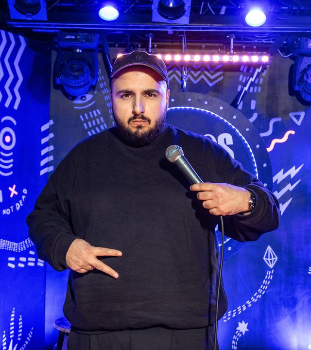 Sezer Oglu steht auf der Bühne und zeigt mit seiner rechten Hand das Peace Zeichen vor seinem Bauch. In der linken Hand hat er ein Mikrofon.