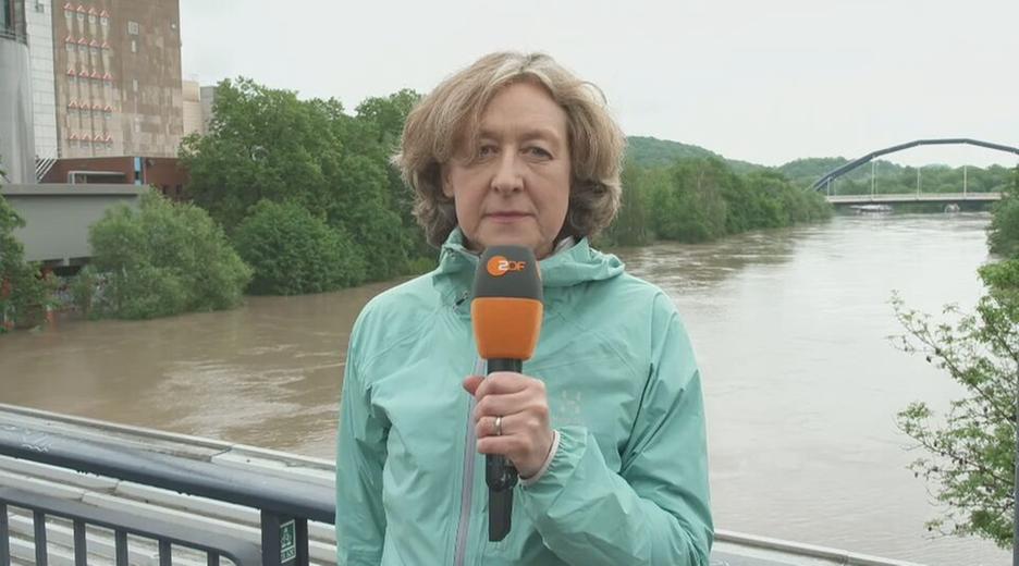 Susanne Freitag-Carteron mit einem ZDF Mikrofon in der Hand auf einer Brücke im Hintergrund ein Fluss.