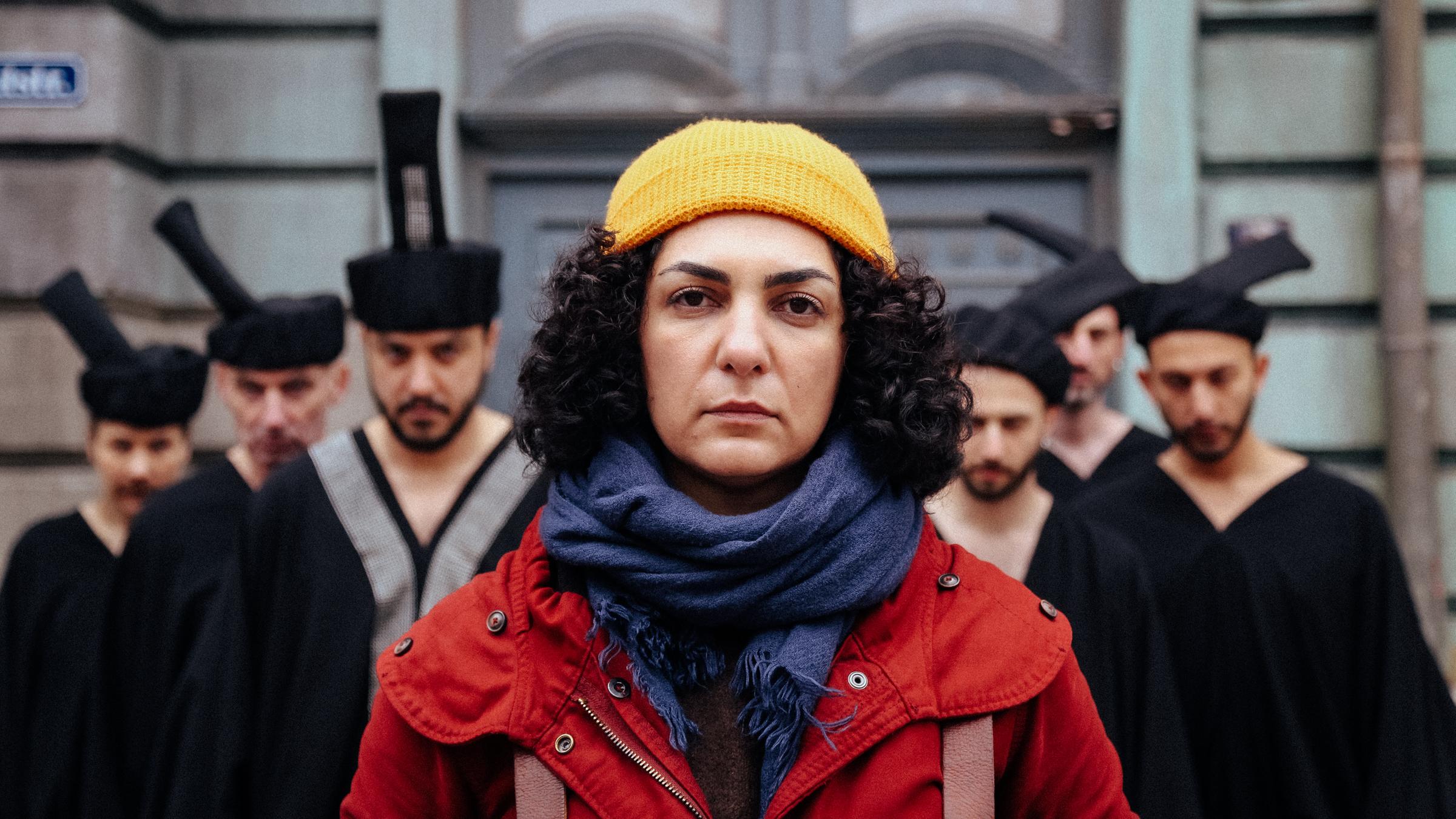"Shahid": Narges (Baharak Abdolifard) steht vor einener Gebäudefassade und blickt in die Kamera. Sie trägt eine gelbe Mütze, ein blaues Halstuch und einen roten Anorak. Im Hintergrund 