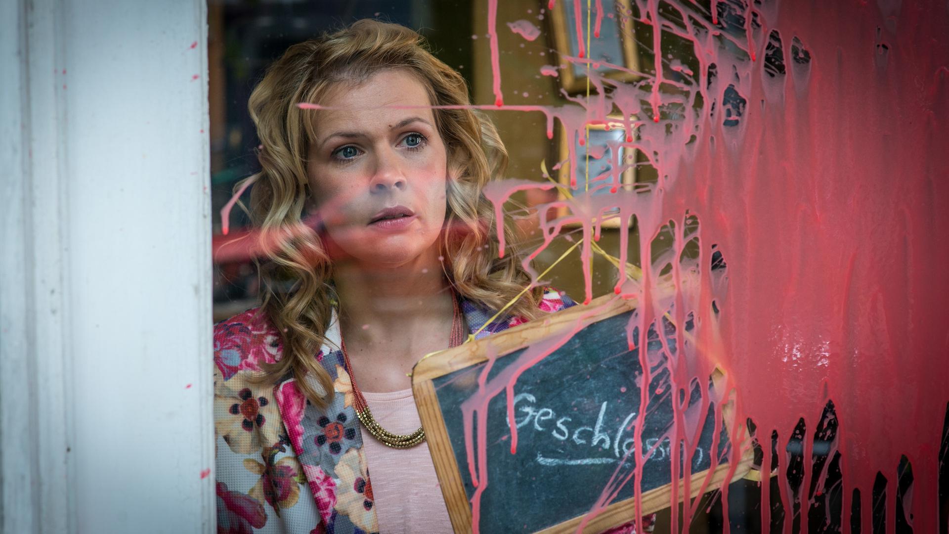 Notruf Hafenkante - Shopping": Nina Hell (Tina Amon Amonsen) steht mit schockiertem Blick hinter einer Fensterscheibe, auf der ein großer Klecks rote Farbe klebt.