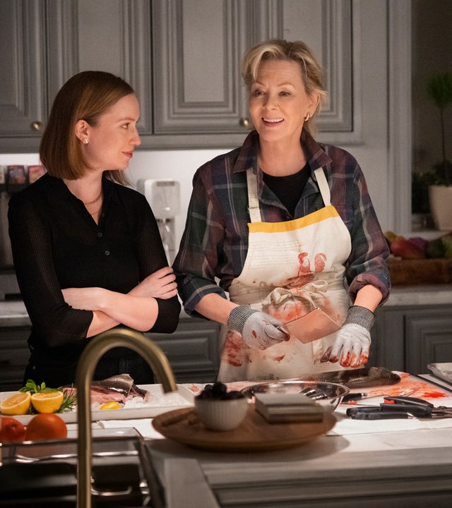 "Hacks - Show, die letzte": Ava (Hannah Einbinder) steht mit verschränkten Armen neben Deborah (Jean Smart) in einer Küche und blickt zu ihr, während Deborah eine blutbefleckte Schürze trägt, mit Handschuhen ein Hackmesser hält und dabei lächelnd in ihre Richtung schaut.