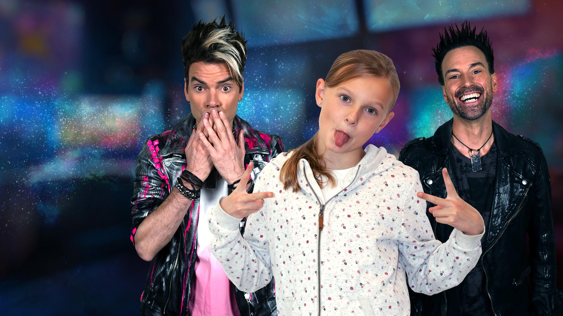 Chris Ehrlich hält sich die Hände vor den Mund. Daneben steht sein Bruder Andreas Ehrlich und lacht. Vor ihnen steht die 10-jährige Ida, die ihre Zunge zur Kamera streckt. Alle drei stehen vor einem bunten Hintergrund.