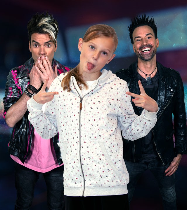 Chris Ehrlich hält sich die Hände vor den Mund. Daneben steht sein Bruder Andreas Ehrlich und lacht. Vor ihnen steht die 10-jährige Ida, die ihre Zunge zur Kamera streckt. Alle drei stehen vor einem bunten Hintergrund.