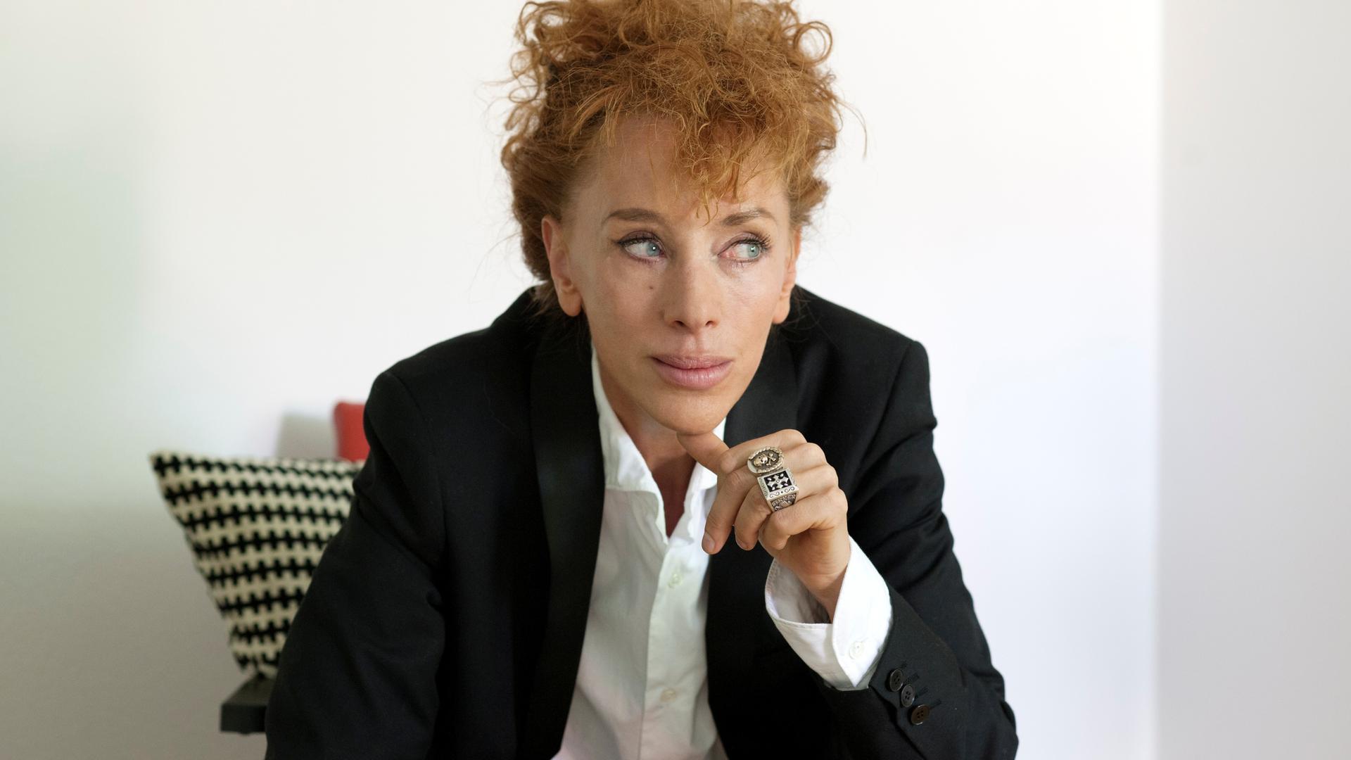 Eine Frau mit wuscheligen roten Haaren hat sich nachdenklich einen Daumen ans Kinn gelegt.