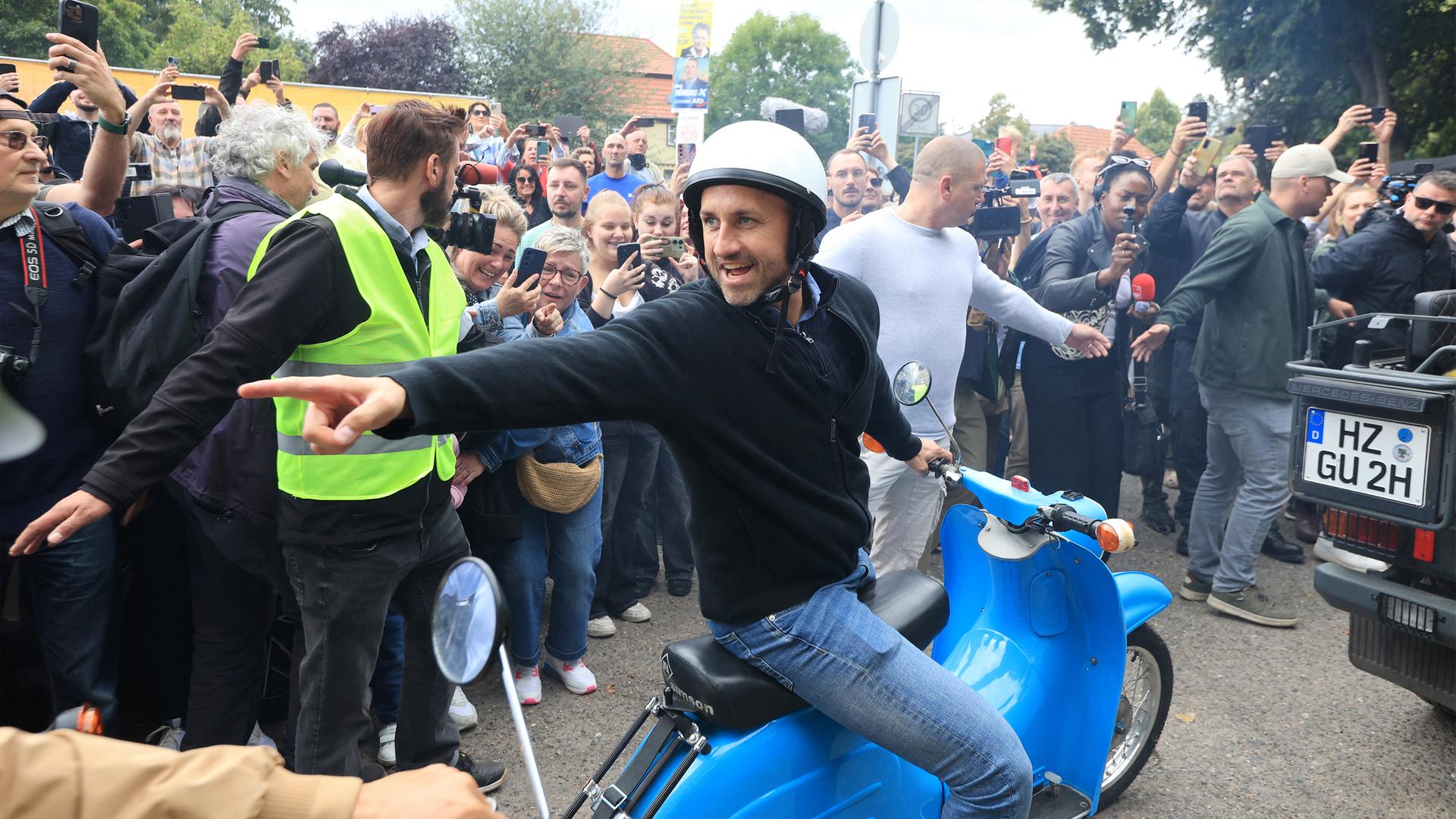 Ein Mann sitzt auf einem blauen Moped und zeigt nach links. Hiter ihm halten zwei Ordner die Menschenmenge davon ab, weiter vor zu treten. 