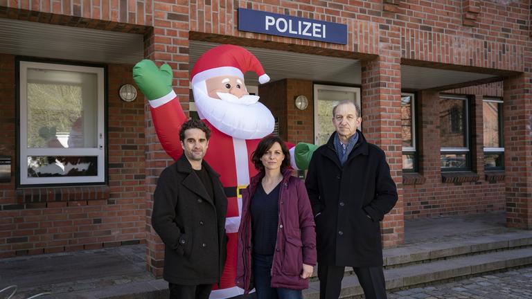 "Nord Nord Mord - Sievers und die Stille Nacht": Hinnerk Feldmann (Oliver Wnuk), Ina Behrendsen (Julia Brendler) und Carl Sievers (Peter Heinrich Brix) stehen vor dem Polizeigebäude. Hinter ihnen steht ein großer aufblasbarer Weihnachtsmann. Das Rotklinkergebäude hat mehrere Fenster. Hinnerk Feldmann trägt einen dunklen Kurzmantel und darunter einen schwarzen Pullover. Ina Behrendsen trägt ihre weinrote halblange Winterjacke offen, darunter einen dunklen V-Ausschnittpullover. Carl Sievers hat die Hände in den Jackentaschen. Seine dunkle Winterjacke ist geschlossen, er trägt einen hellblauen Schal. Alle drei schauen geradeaus in die Kamera.