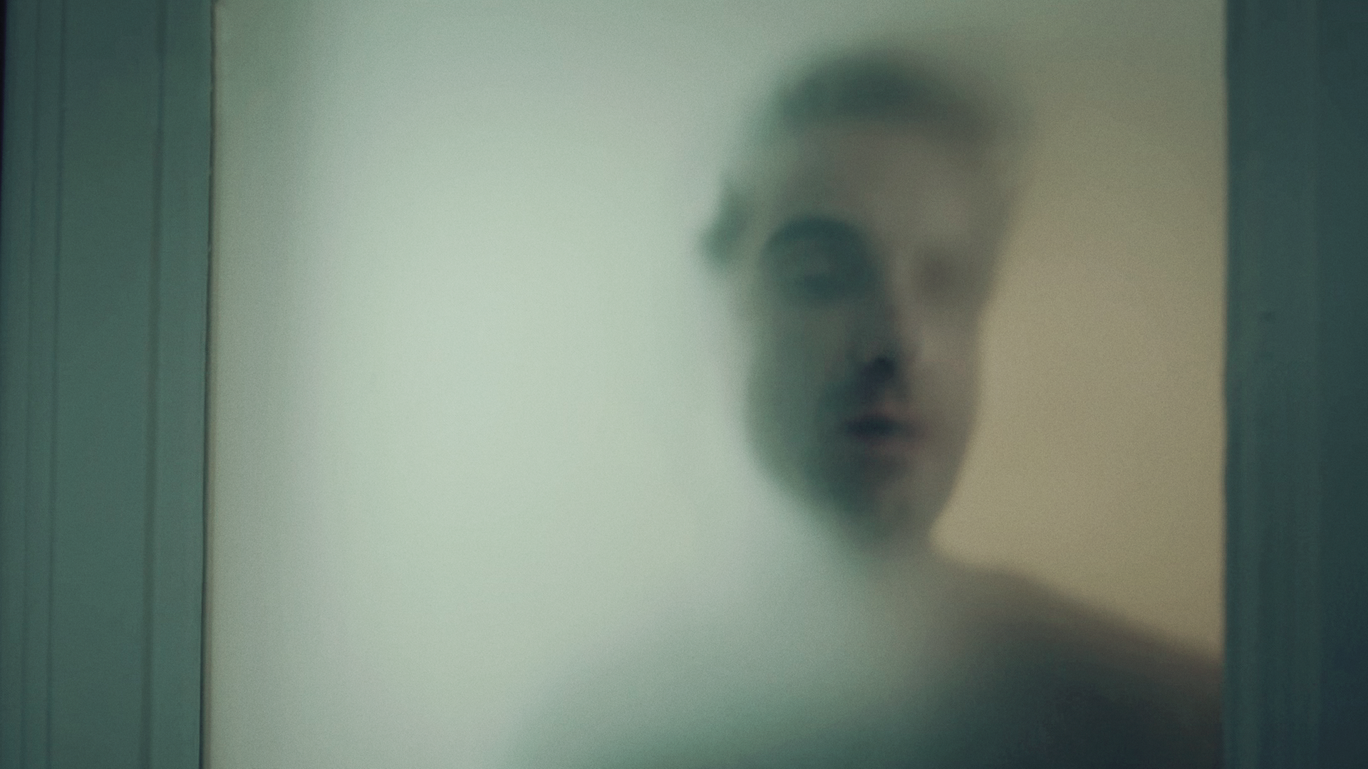 Hinnerk Feldmann (Oliver Wnuk) steht hinter einer Milchglasscheibe, sein Gesicht und sein Körper sind nur schemenhaft zu erkennen. 