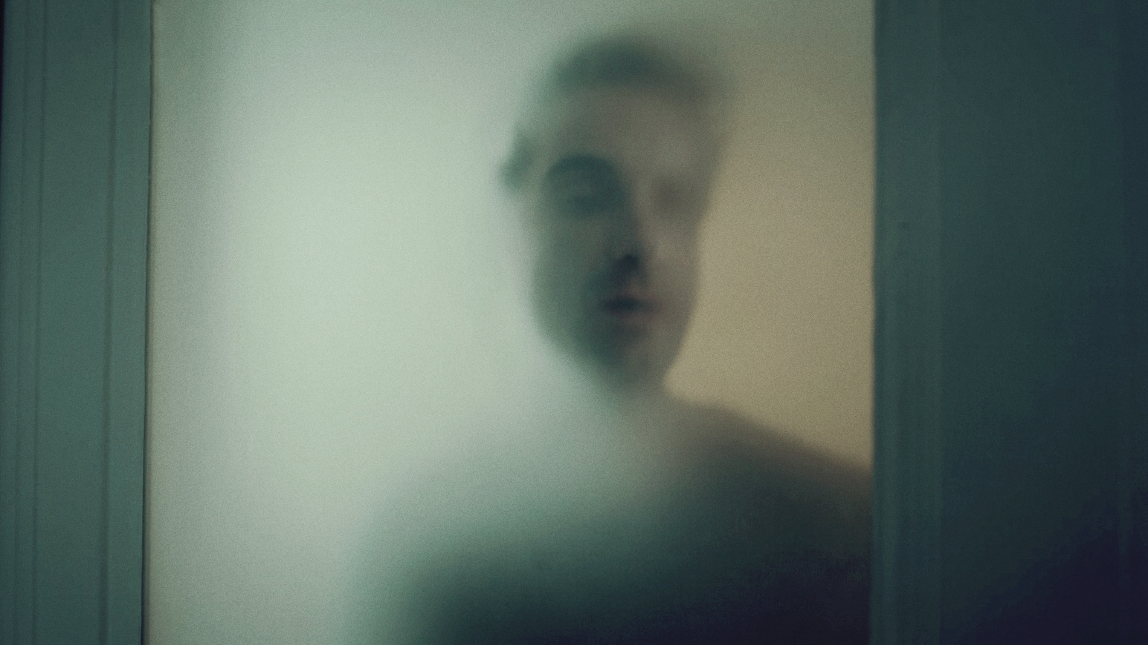 Hinnerk Feldmann (Oliver Wnuk) steht hinter einer Milchglasscheibe, sein Gesicht und sein Körper sind nur schemenhaft zu erkennen. 