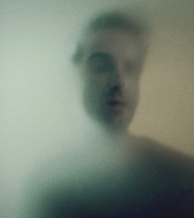 Hinnerk Feldmann (Oliver Wnuk) steht hinter einer Milchglasscheibe, sein Gesicht und sein Körper sind nur schemenhaft zu erkennen. 