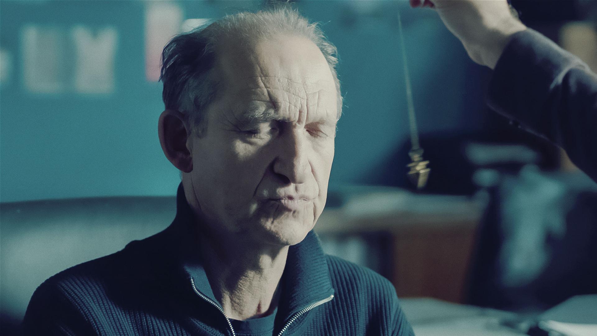 Carl Sievers (Peter Heinrich Brix) sitzt mit geschlossenen Augen auf einem Stuhl und vor seinem Gesicht baumelt ein Pendel.