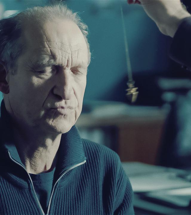 Carl Sievers (Peter Heinrich Brix) sitzt mit geschlossenen Augen auf einem Stuhl und vor seinem Gesicht baumelt ein Pendel.
