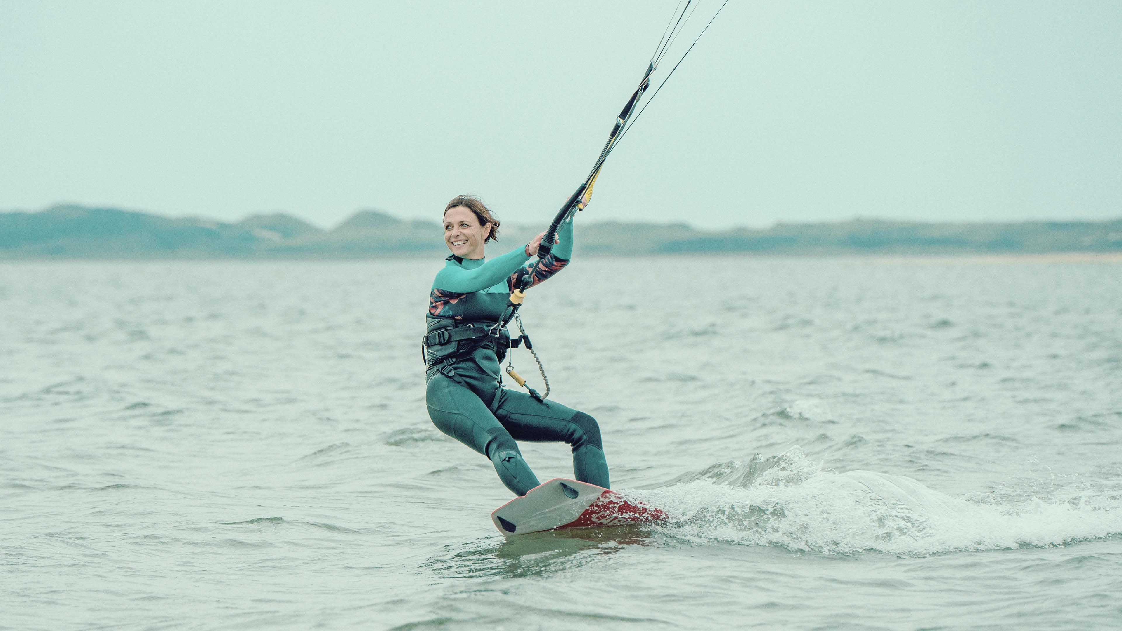 Ina Behrendsen (Julia Brendler) steht auf einem Surfboard im Wasser und wird von einem Lenkdrachen gezogen. Sie trägt einen grünen Neoprenanzug mit türkis-farbenem Einsatz an Armen und Hals. Das Meer ist grau und der Himmel trüb.