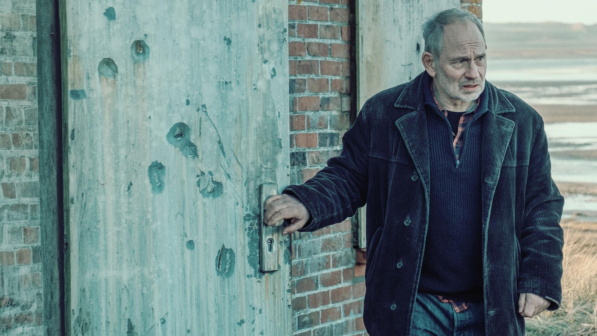 Siggi (Michael Lott) steht an einem roten Backsteinhäuschen im Watt. Er sieht ernst nach vorne, während er eine schwere Holztür öffnet. Er trägt Jeans und eine dunkelbraune Cordjacke.