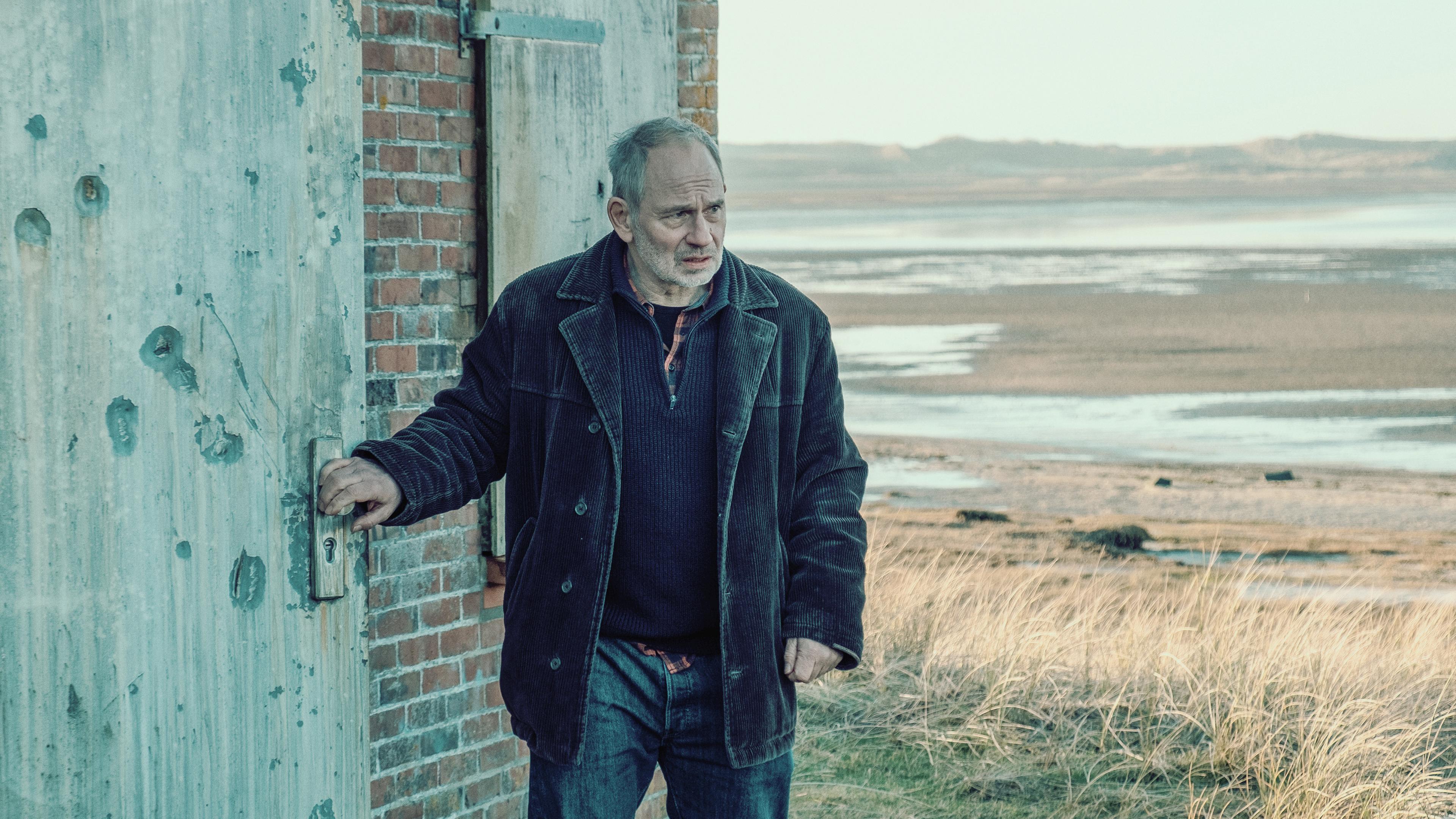 Siggi (Michael Lott) steht an einem roten Backsteinhäuschen im Watt. Er sieht ernst nach vorne, während er eine schwere Holztür öffnet. Er trägt Jeans und eine dunkelbraune Cordjacke.