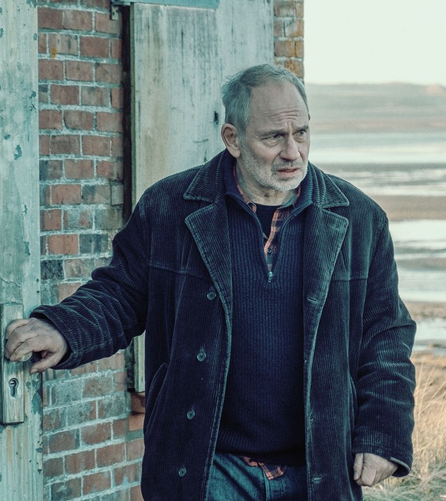 Siggi (Michael Lott) steht an einem roten Backsteinhäuschen im Watt. Er sieht ernst nach vorne, während er eine schwere Holztür öffnet. Er trägt Jeans und eine dunkelbraune Cordjacke.