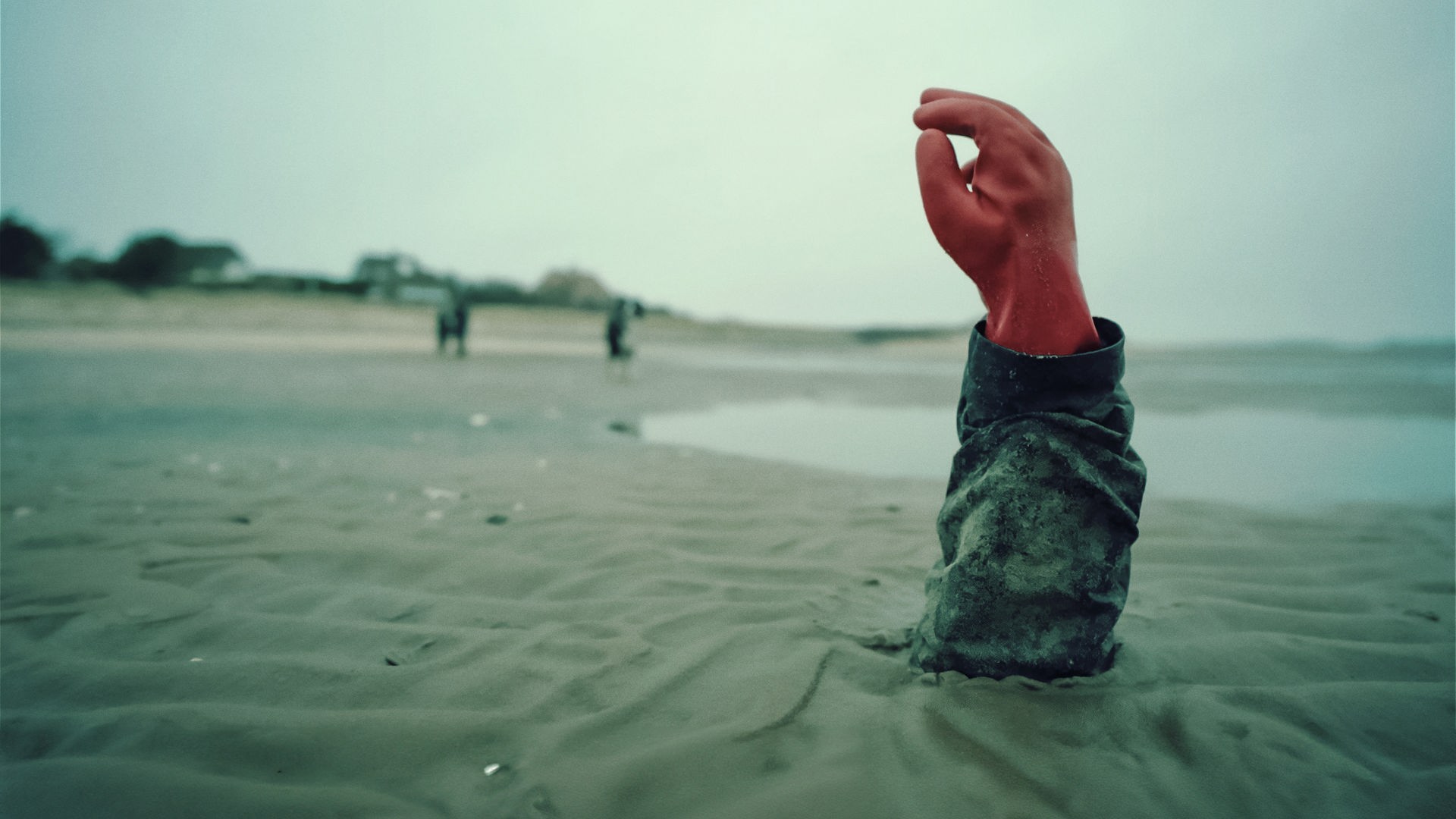 Ein grauer Sandstrand. Aus dem Watt ragen Arm und Hand eines Menschen. Die Hand ist mit einem roten Gummihandschuh bekleidet. Im Hintergrund schemenhaft zu erkennen: vereinzelte Häuser, drei Personen.