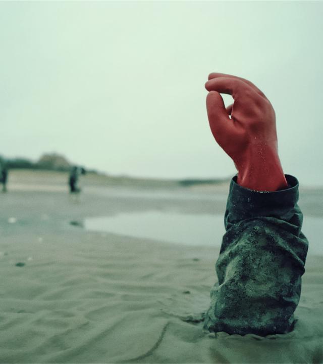 Ein grauer Sandstrand. Aus dem Watt ragen Arm und Hand eines Menschen. Die Hand ist mit einem roten Gummihandschuh bekleidet. Im Hintergrund schemenhaft zu erkennen: vereinzelte Häuser, drei Personen.