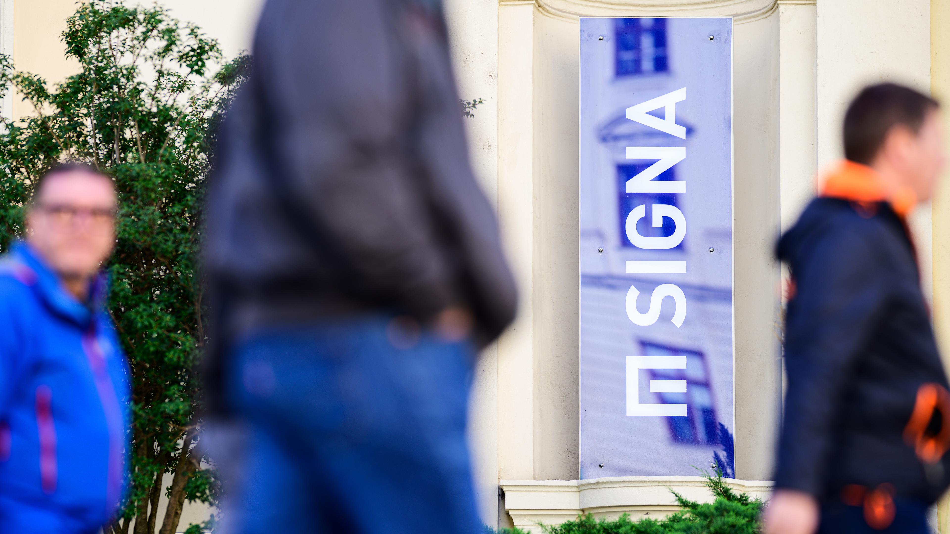 Menschen gehen an einem Logo des österreichischen Immobilien- und Handelsunternehmens Signa Holding im Wiener Büro der Gruppe vorbei, aufgenommen am 08.11.2023