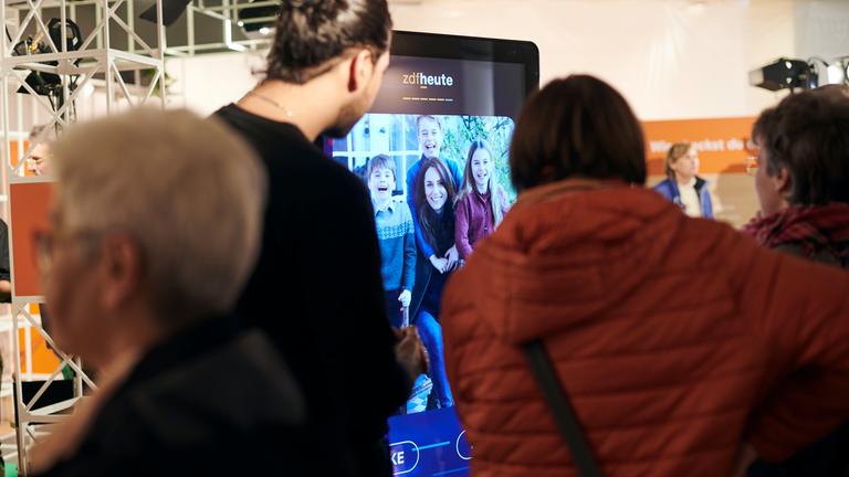 Das ZDF auf dem Silbersalz Festival 25: Von hinten aufgenommen: Zuschauer vor einer Lichtbildwand, auf dem die Besucher den "Fake-Checker" des ZDF ausprobieren können