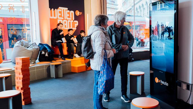 Ein großer Bildschirm mit dem ZDFheute-Logo steht rechts am ZDF-Stand auf dem Silbersalz Festival 2026 in Halle (Saale). Davor stehen zwei Besucher nebeneinander. Links befinden sich Sitzsäcke, eine orangefarbene Sitzplattform und ein Turm aus Holzklötzen. Drei weitere Personen sitzen vor einer schwarzen Wand mit orangefarbener Schrift und ZDF-Logo. Große Fenster geben den Blick auf die Straße frei