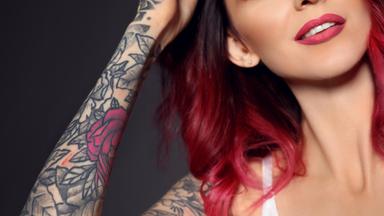 Nahaufnahme einer jungen Frau vor dunklem Hintergrund. Sie trägt einen Nasenring und ein weißes Oberteil. Ihr angehobener Arm ist großflächig tätowiert; zwischen schwarzen Blumen- und Linienmustern ist eine rote Rose zu sehen. Ihre Hand liegt auf dem Kopf