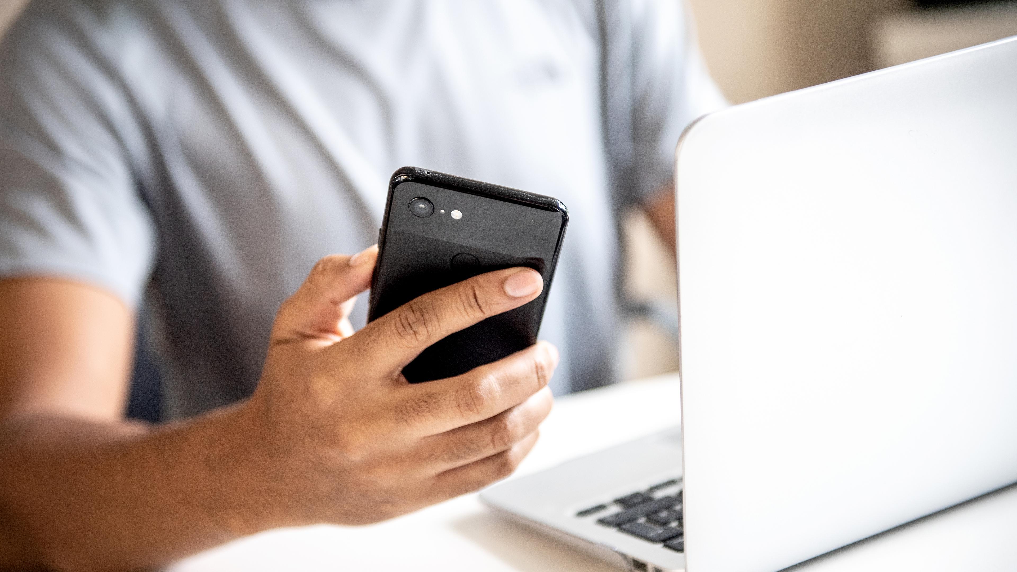 Ein Mann sitzt vor einem Laptop und hält ein schwarzes Smartphone in der rechten Hand.